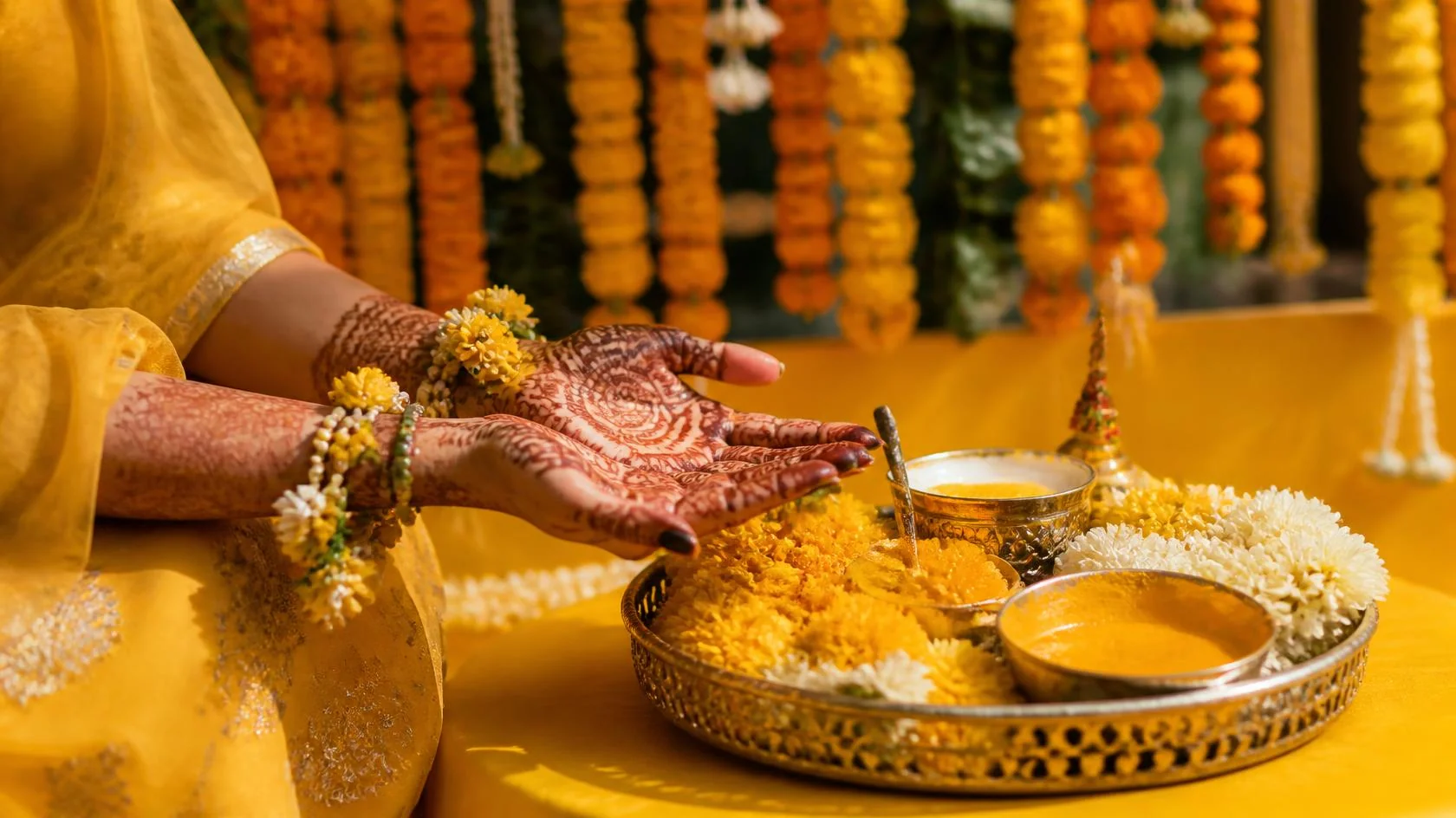 Mehendi-covered hands beside a brass haldi thali with turmeric paste and marigold flowers
