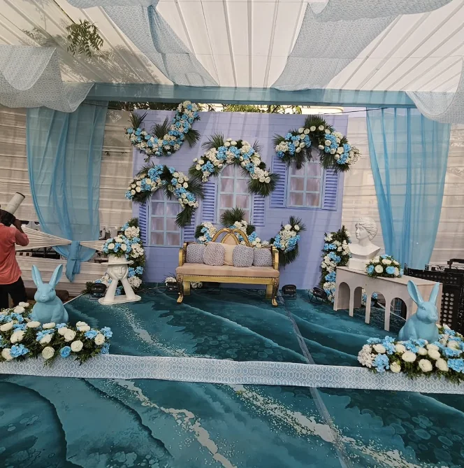 Blue and white carnival-style Haldi decor with floral backdrop, draped canopy, statement sofa and playful themed props